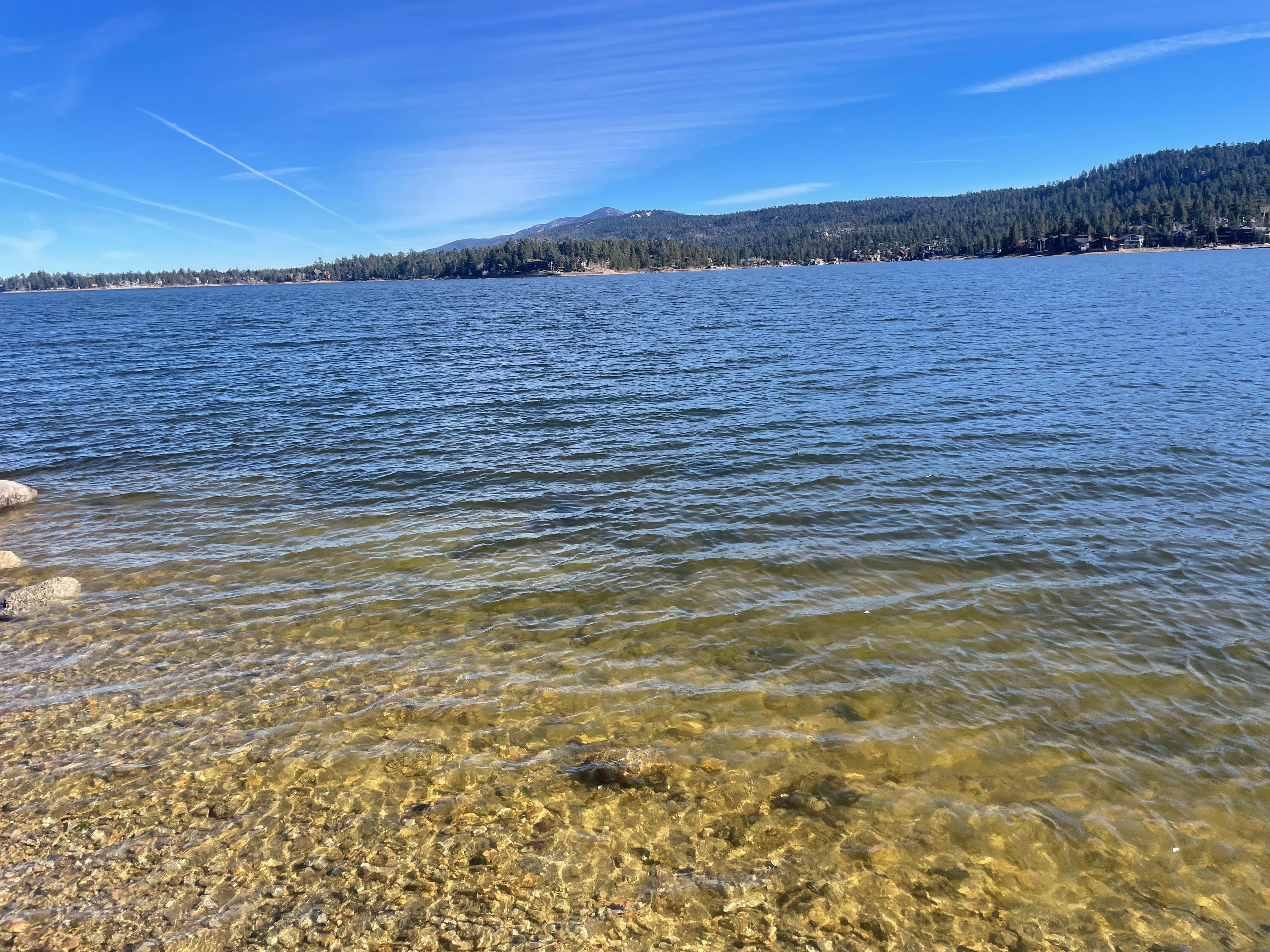 Clear, shallow water over golden rocks at the edge of Big Bear Lake, with pine-covered hills and a mountain peak across the lake