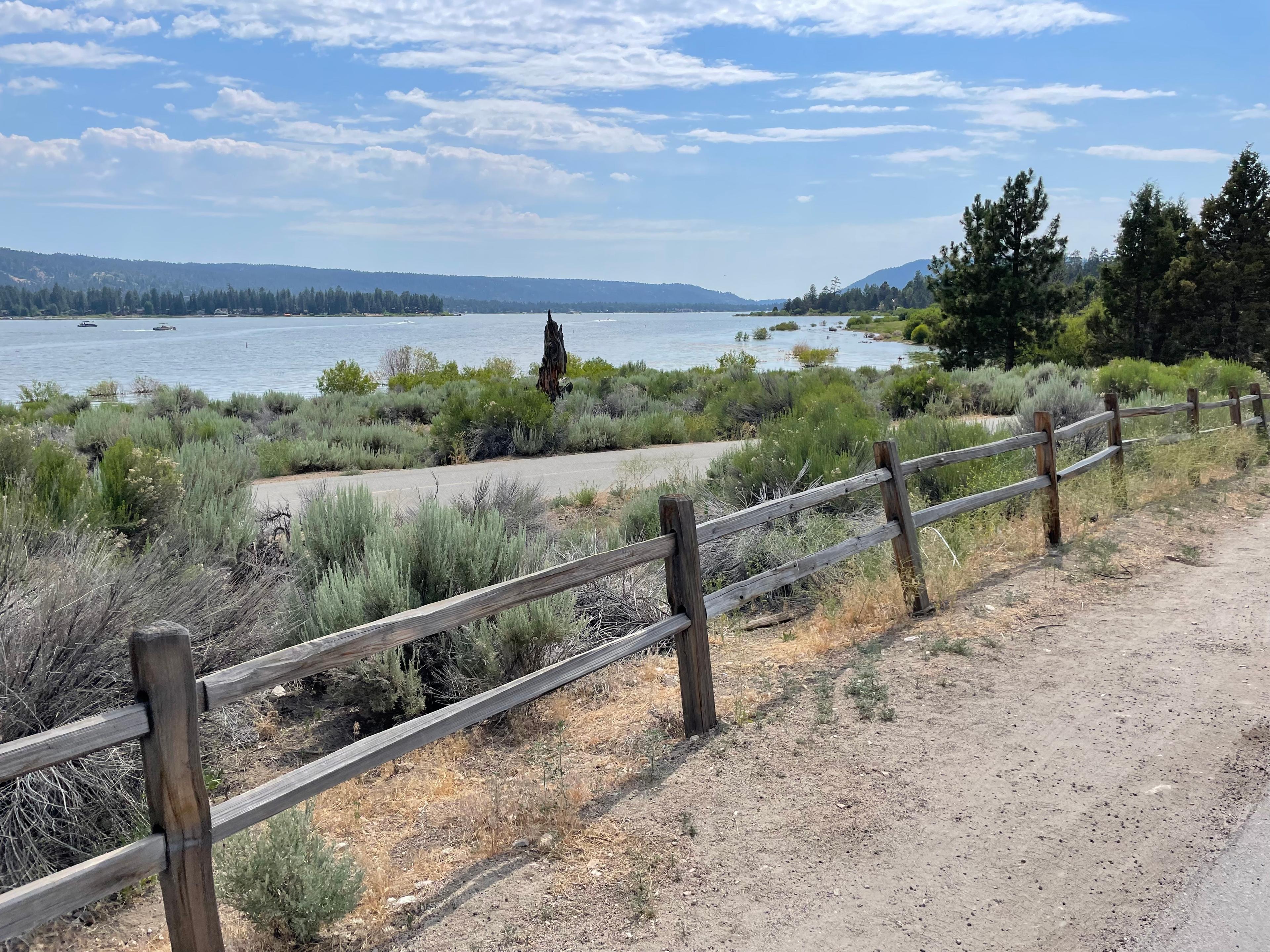 Big Bear Lake from a lakeside trail, with a split-rail fence and sagebrush in front and pine-covered hills along the far shore
