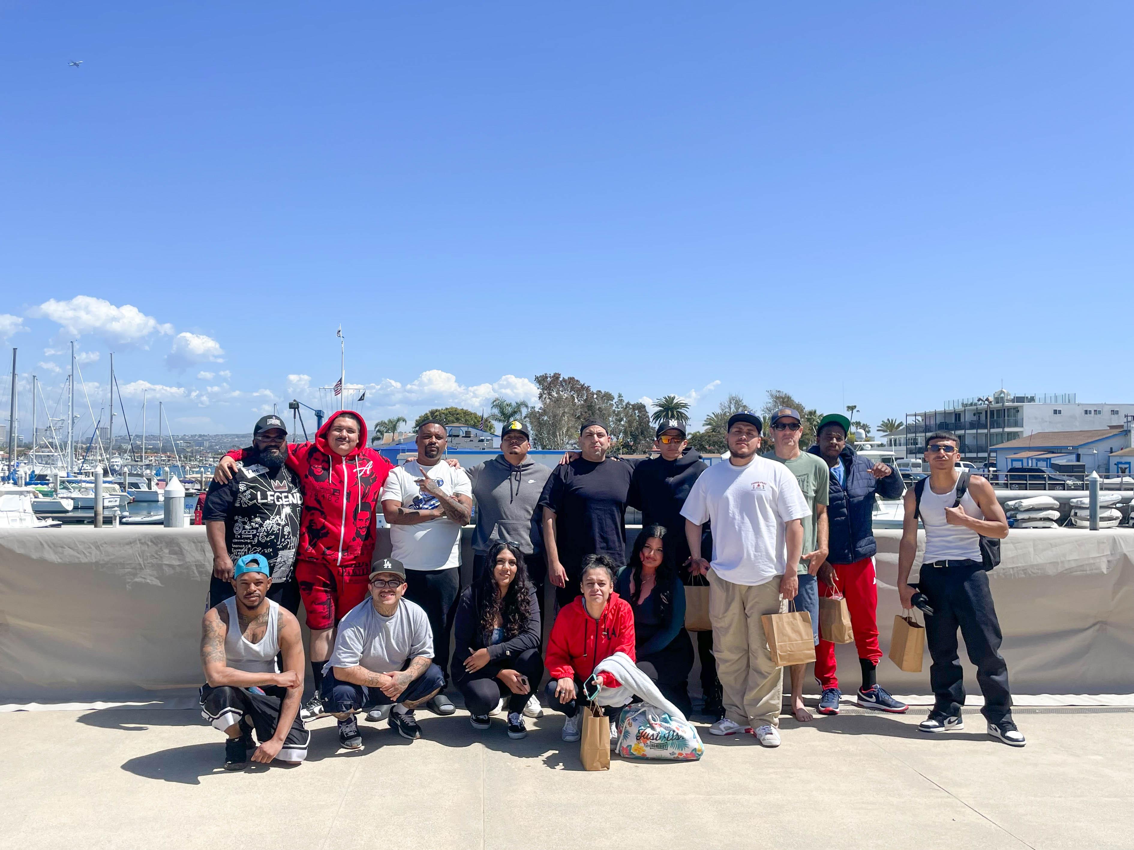 A large group of Outside the City participants gathered on the boardwalk at Marina Park in Newport Beach, with the harbor behind them