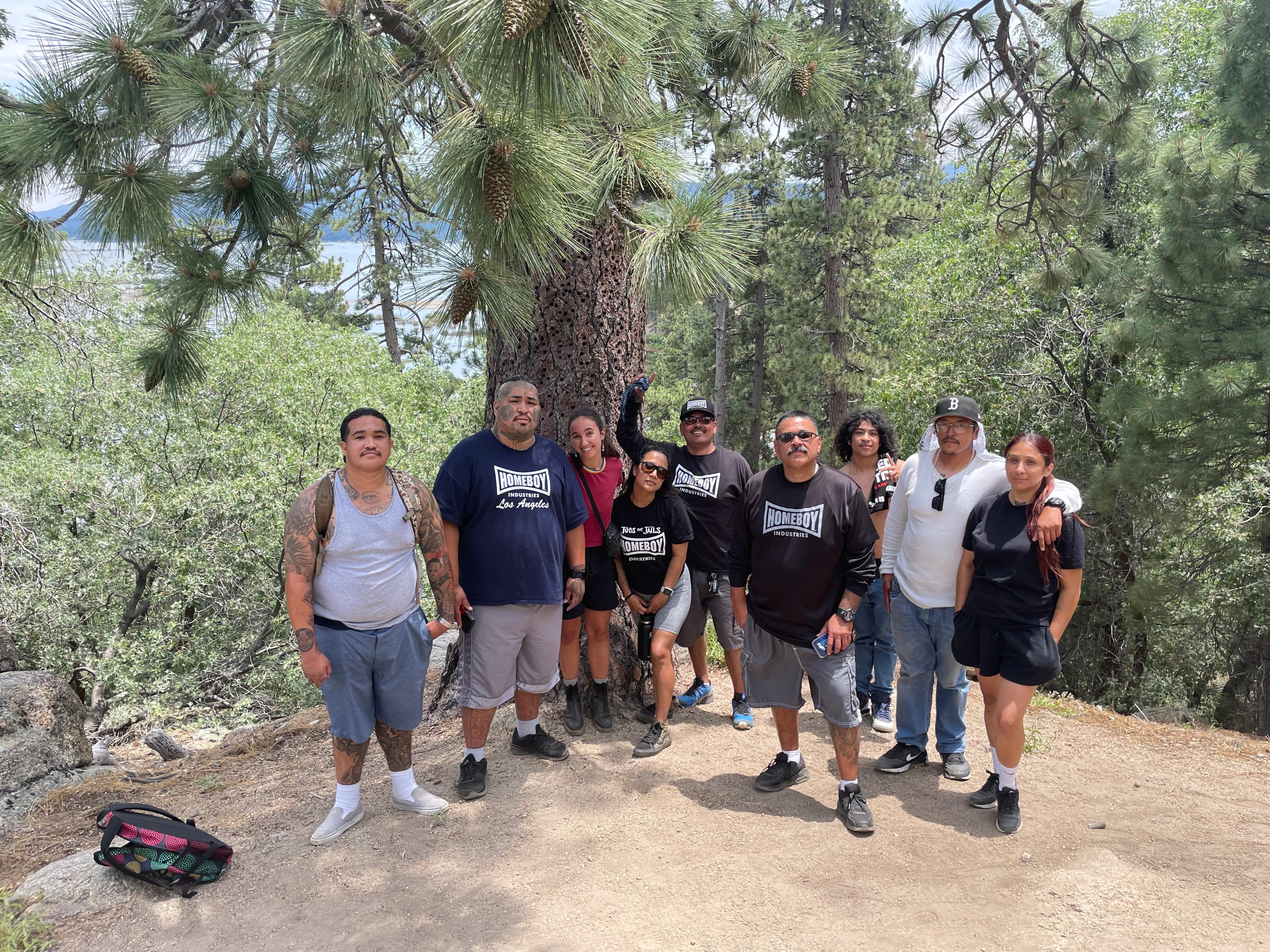 A group of Outside the City participants standing together on a forest trail under tall pines, with Big Bear Lake behind them