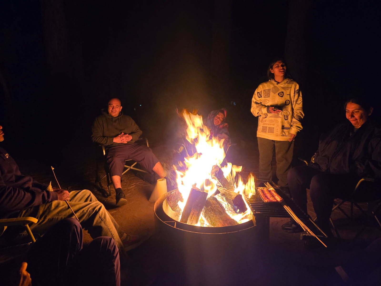 Campers gathered around a blazing fire pit at night, some in camp chairs and one standing and laughing