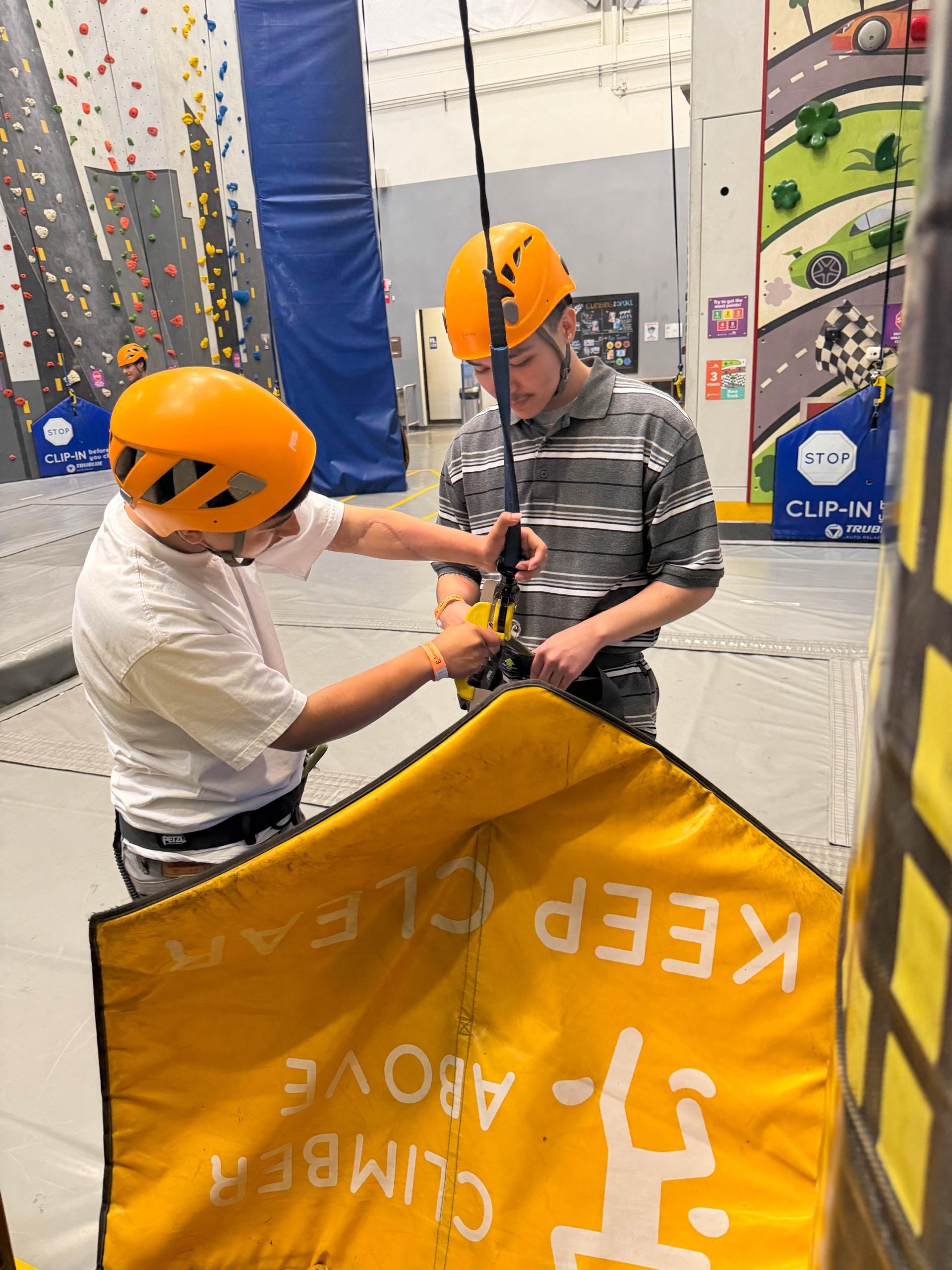 Two participants in orange helmets clip into an auto-belay rope at an indoor climbing gym, beside a yellow safety pad