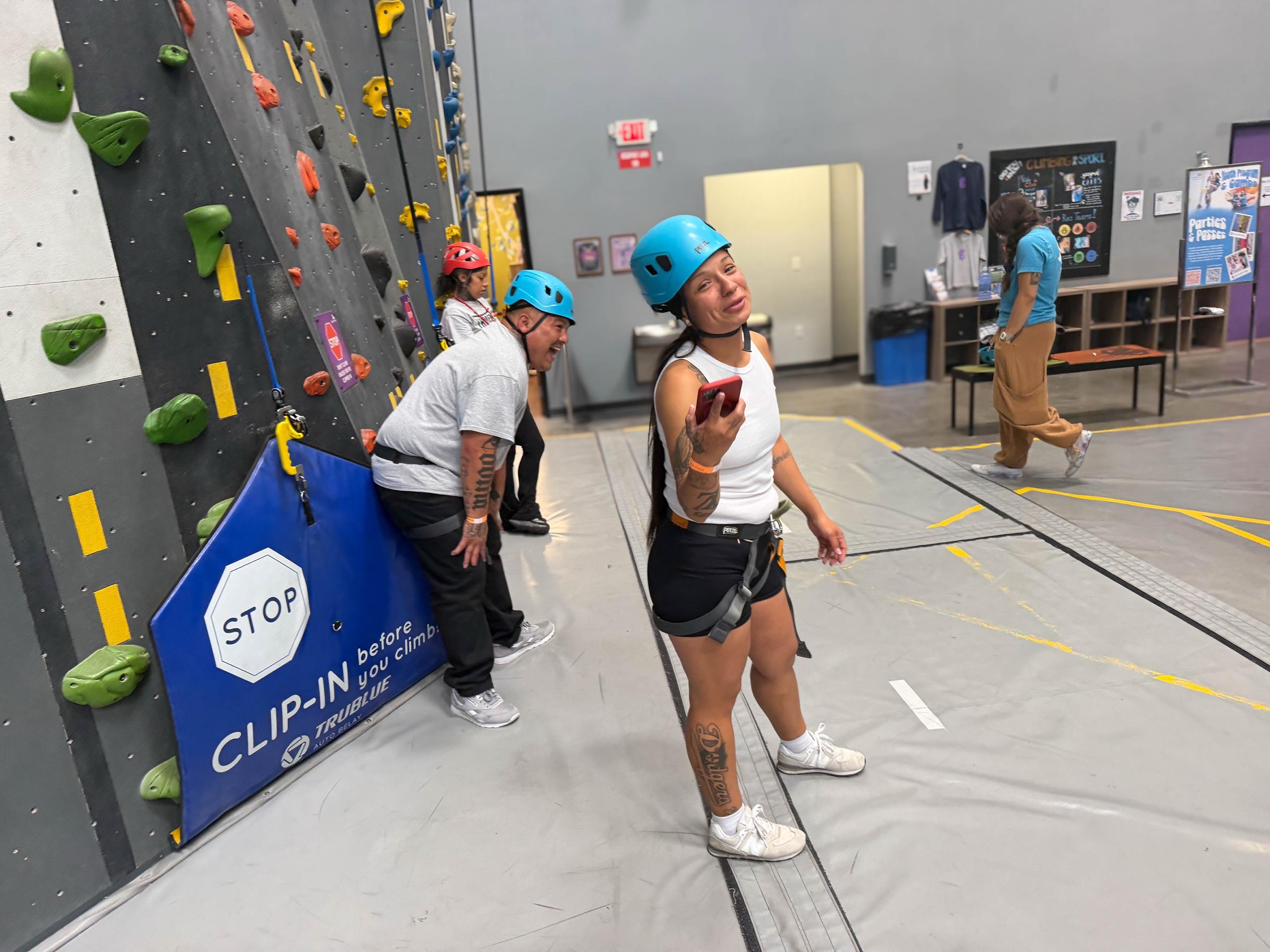 Participants in blue helmets and harnesses laugh together at the base of an indoor climbing wall