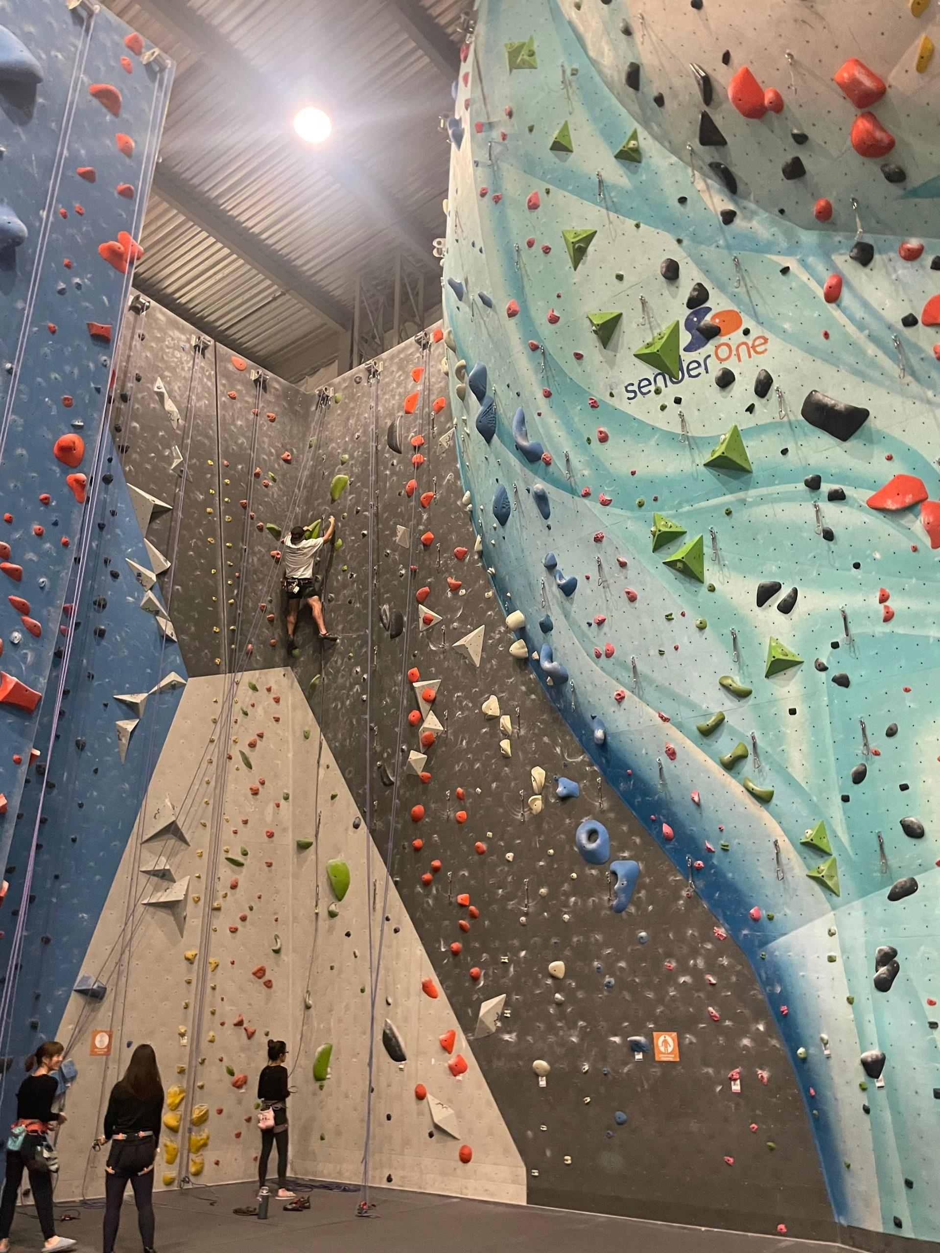 A climber on a rope high up a towering indoor climbing wall at Sender One, with climbers in harnesses watching from the floor