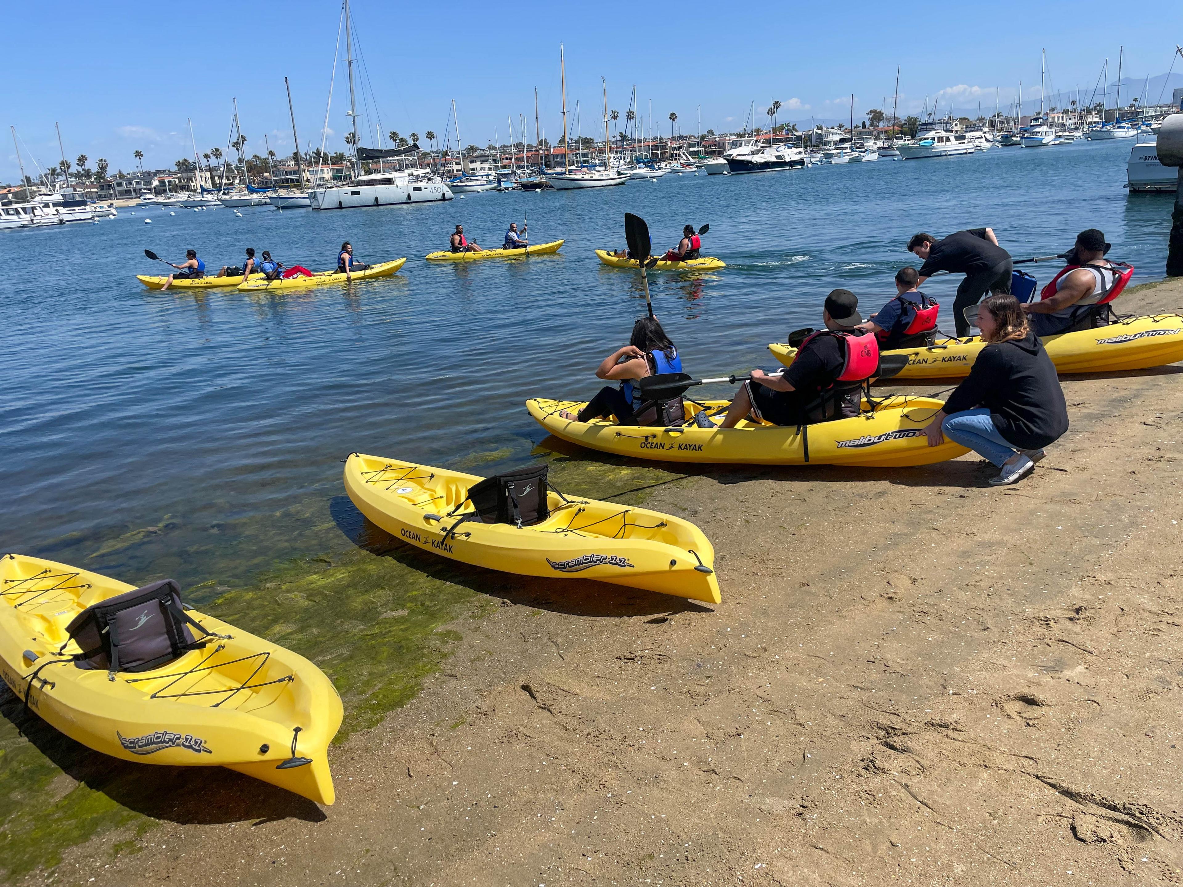 Participants in life vests launching yellow kayaks from a sandy beach on Newport Harbor, with others already paddling past moored sailboats