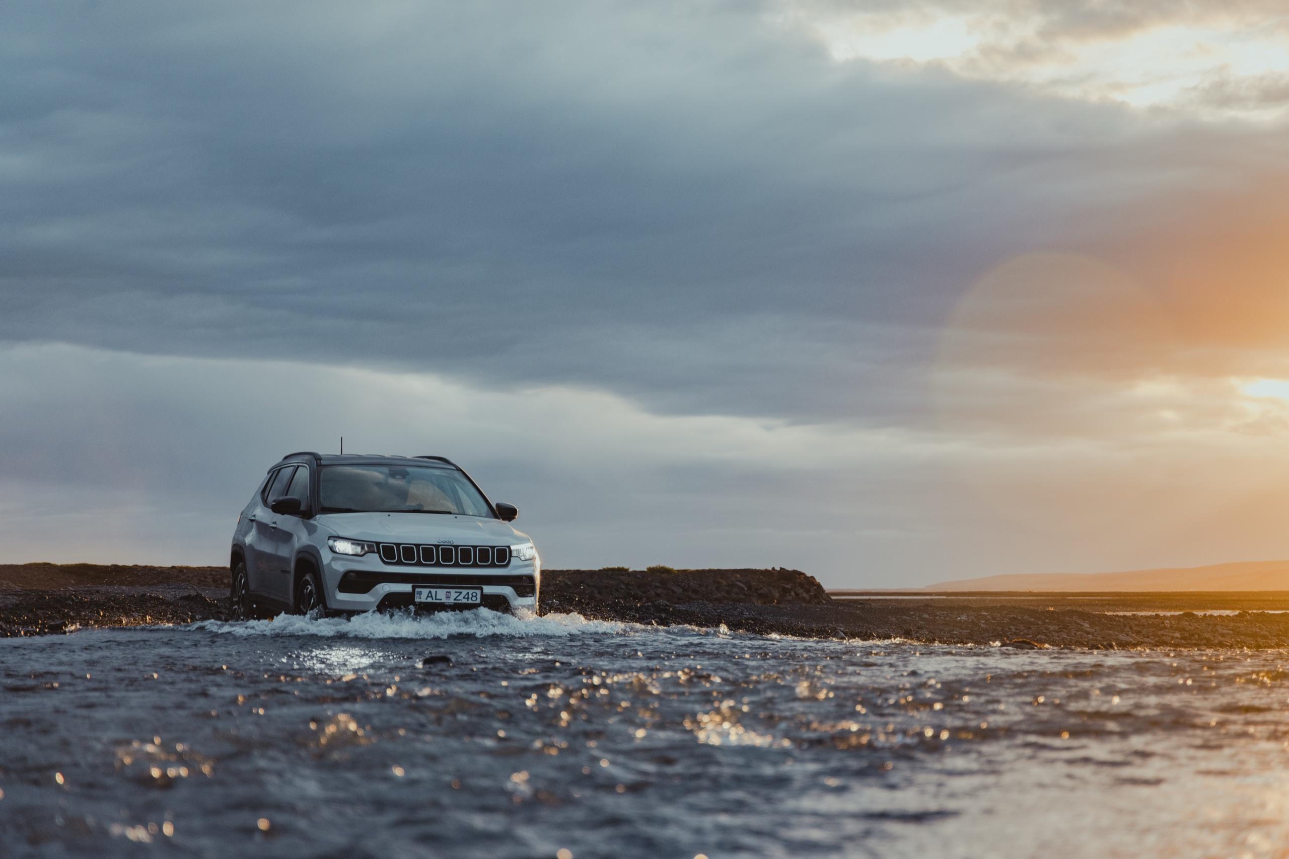 A white Jeep Compass fords a shallow river over black gravel in Iceland, under storm clouds lit by the setting sun