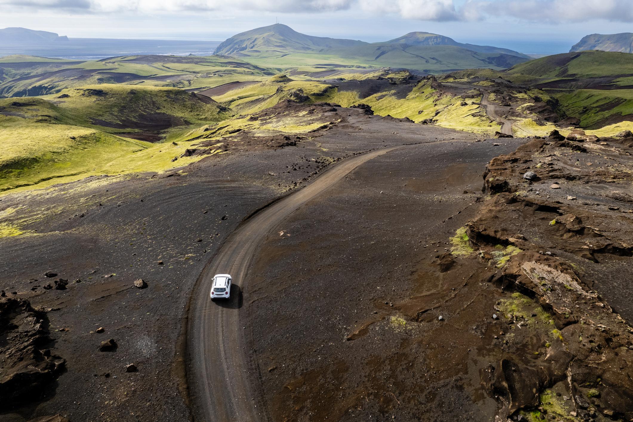 Aerial view of a white SUV on a winding gravel road through black volcanic sand and green mossy hills on Iceland's south coast, with the ocean in the distance