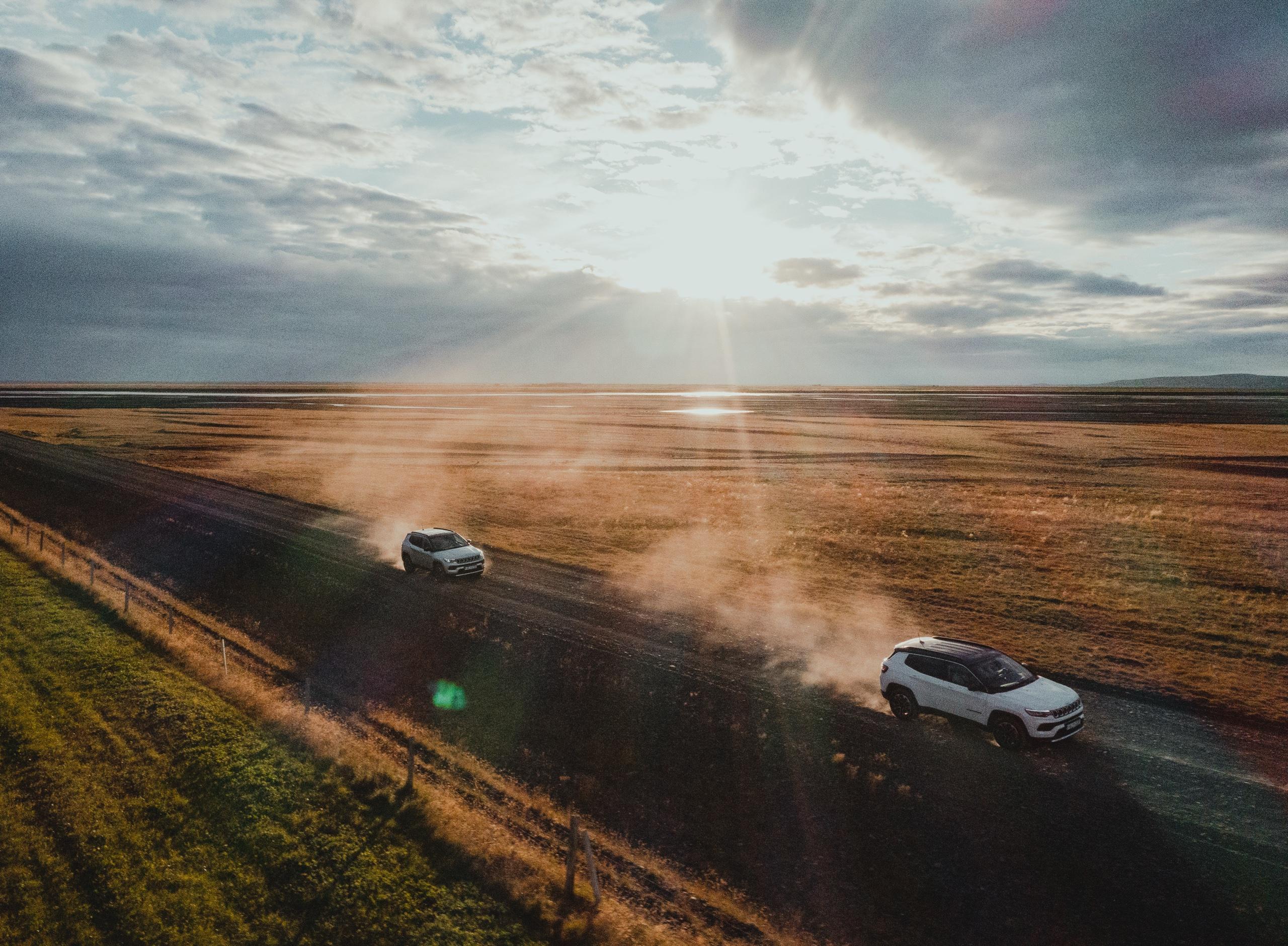 Two white SUVs kick up dust on a gravel road across open grassland in Iceland, with the sun breaking through clouds ahead