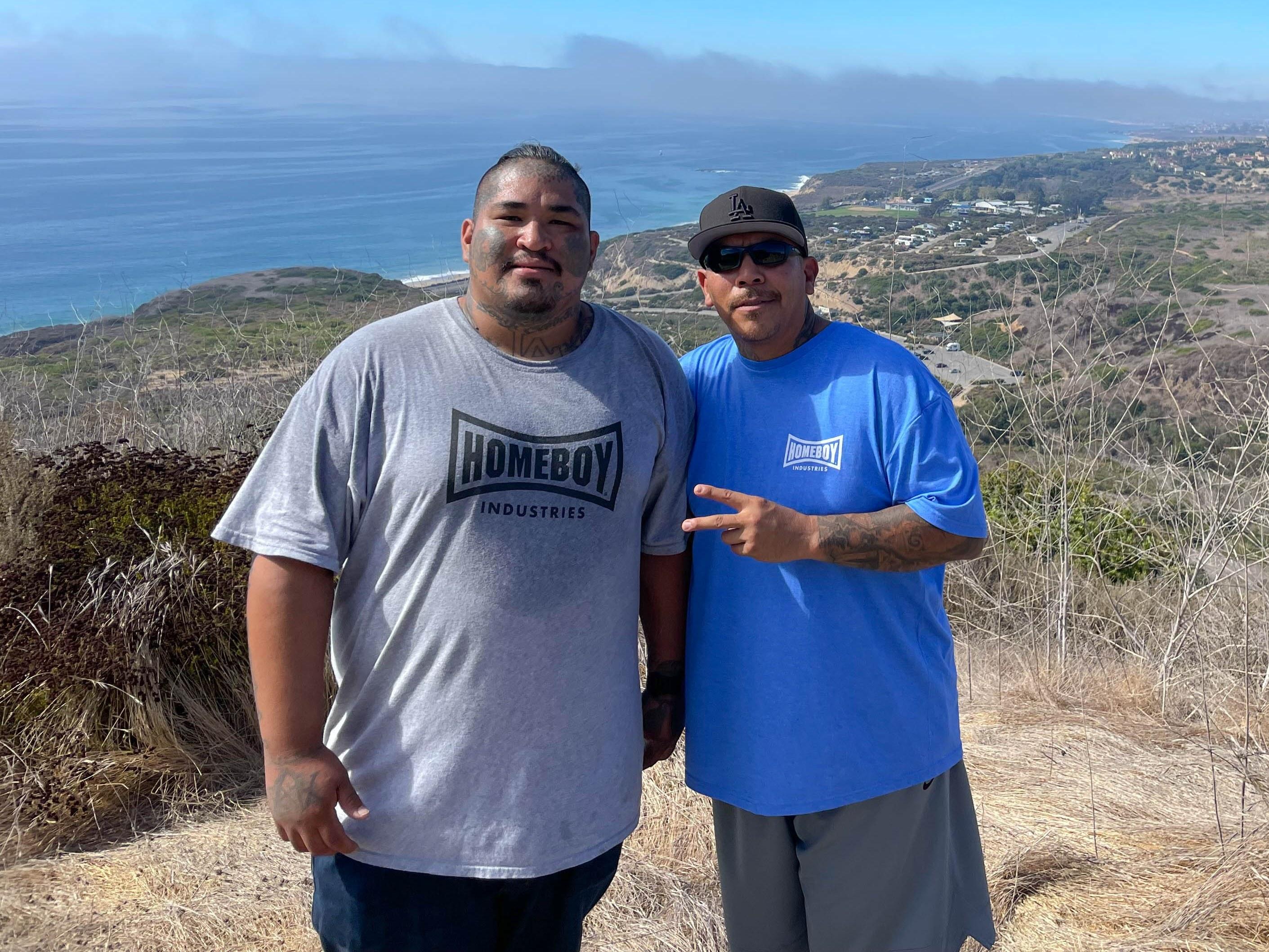 Two participants in Homeboy Industries shirts pose on a hilltop at Crystal Cove State Park, with the coastline and ocean stretching out behind them