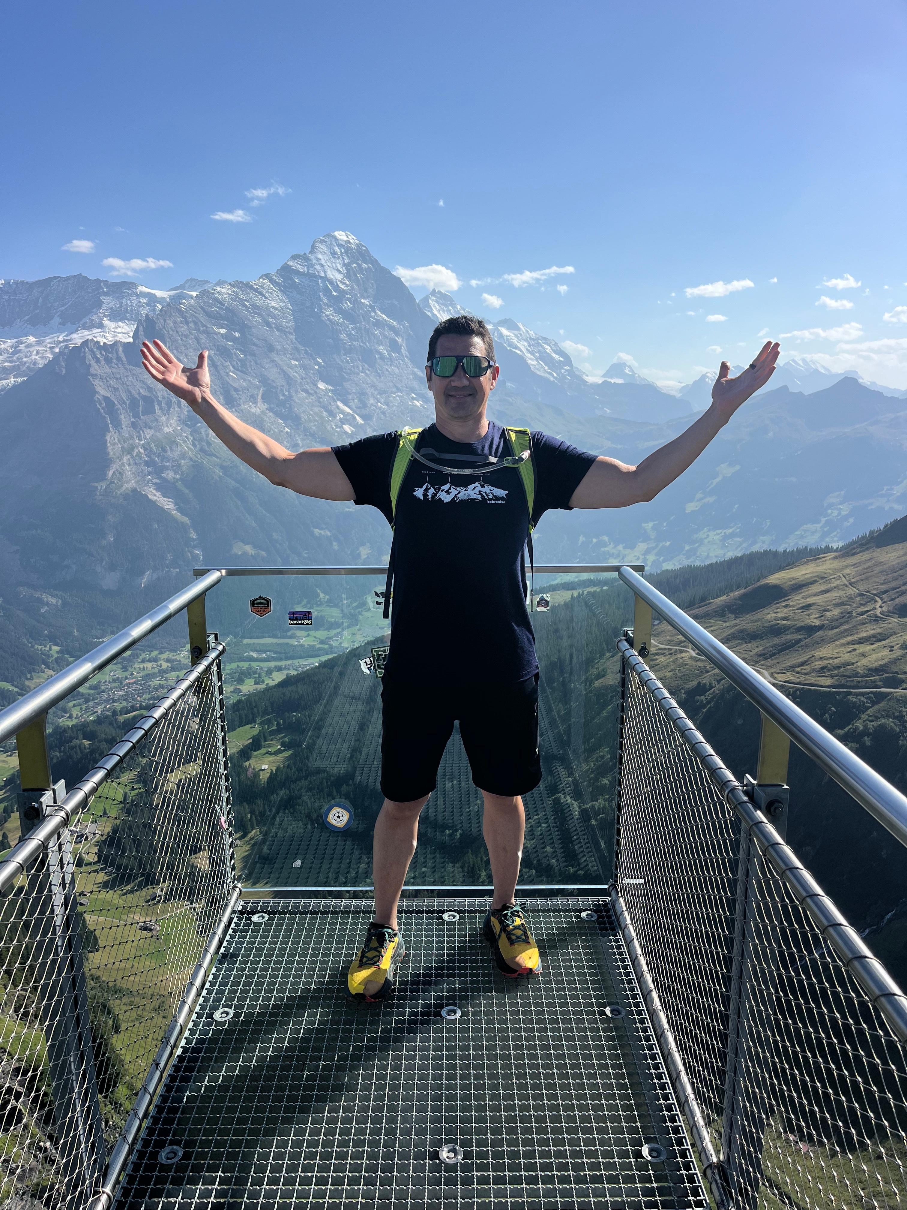 A hiker stands with arms raised on a railed overlook platform, with mountain peaks behind