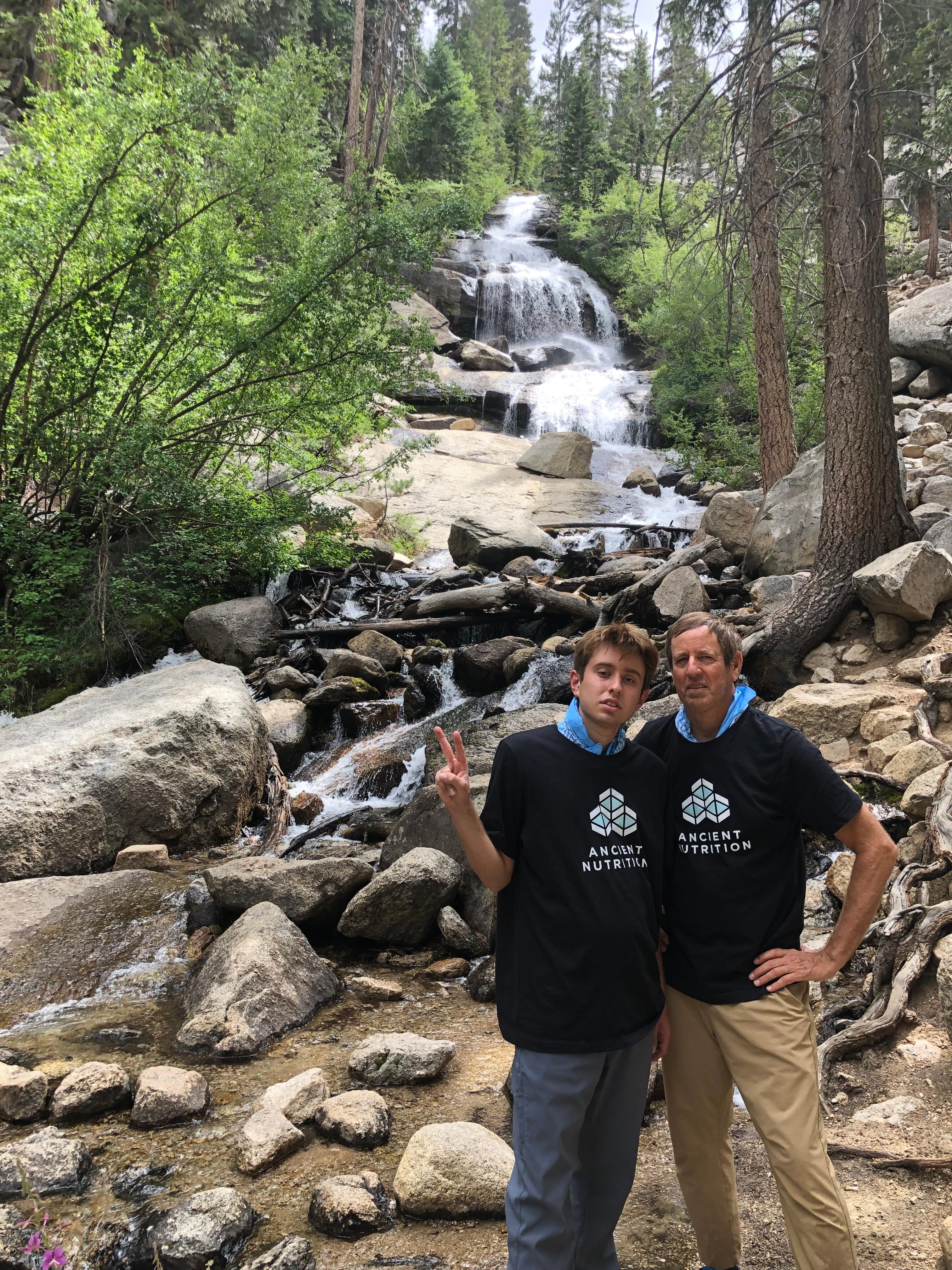 Two hikers pose in front of a waterfall tumbling over boulders in a pine forest