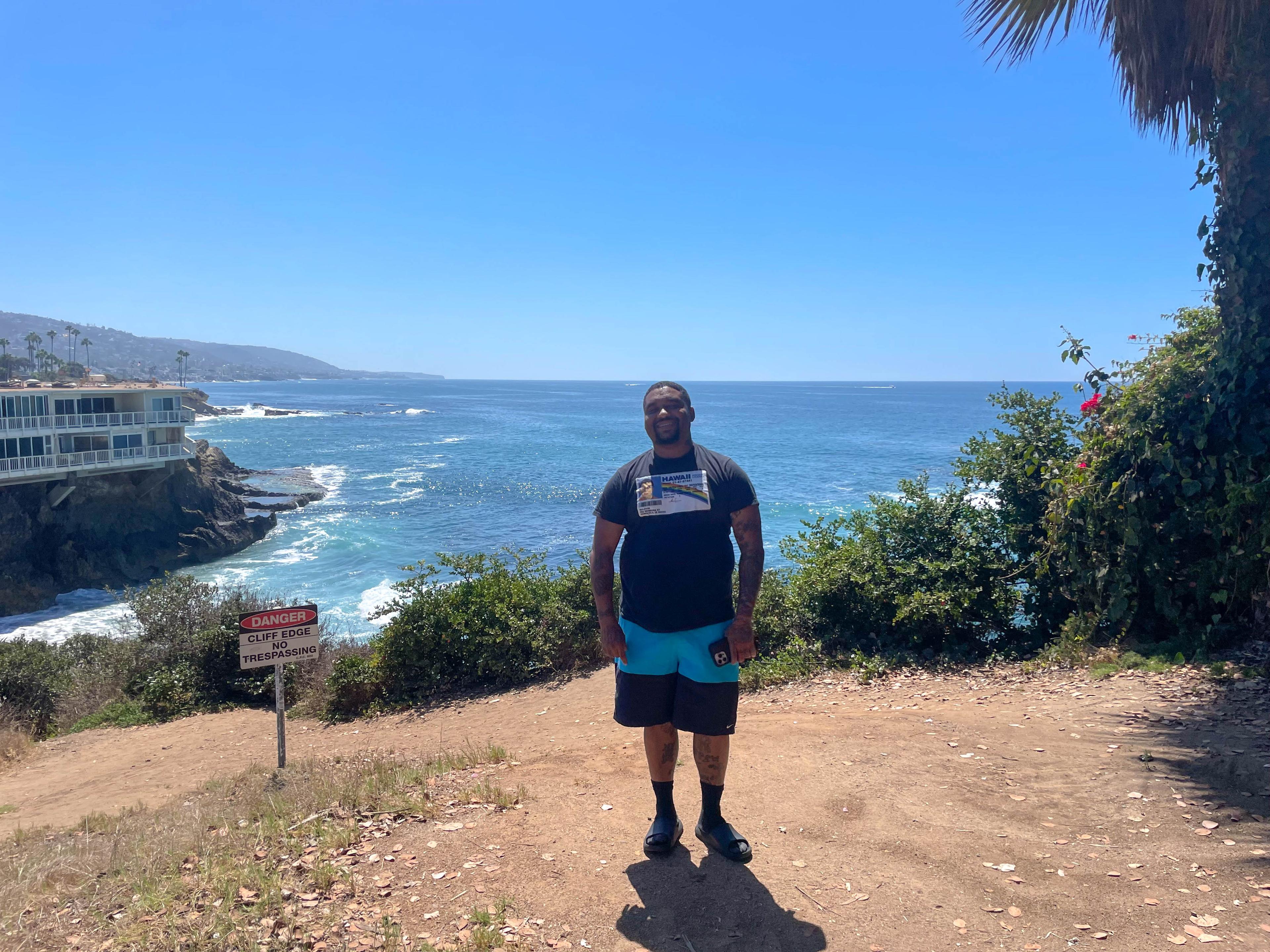 A smiling hiker stands on a clifftop trail in Laguna Beach, with waves breaking on the rocky coves below