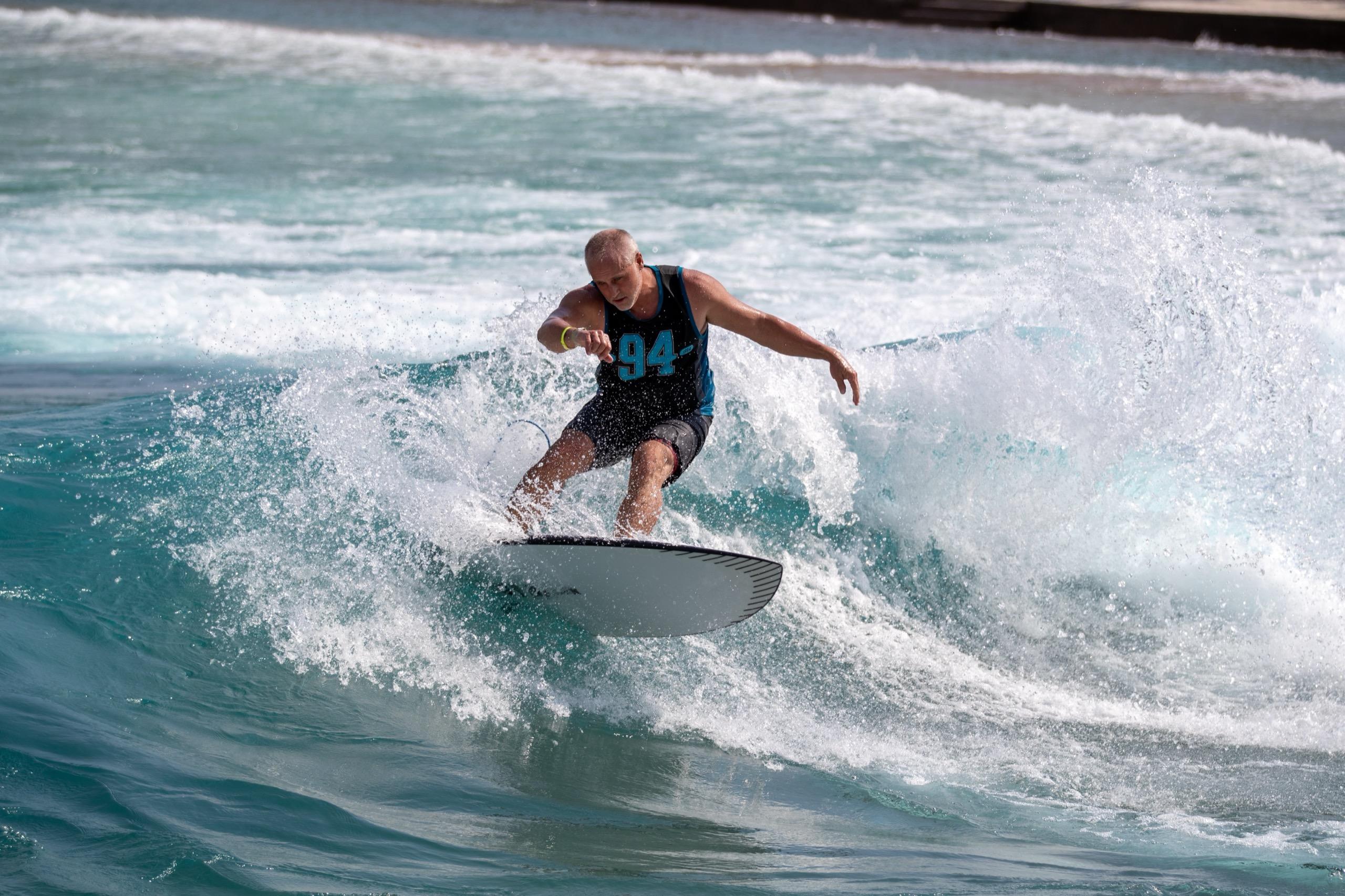 A surfer in a black tank top carves across a breaking wave at a surf park, spray flying around the board