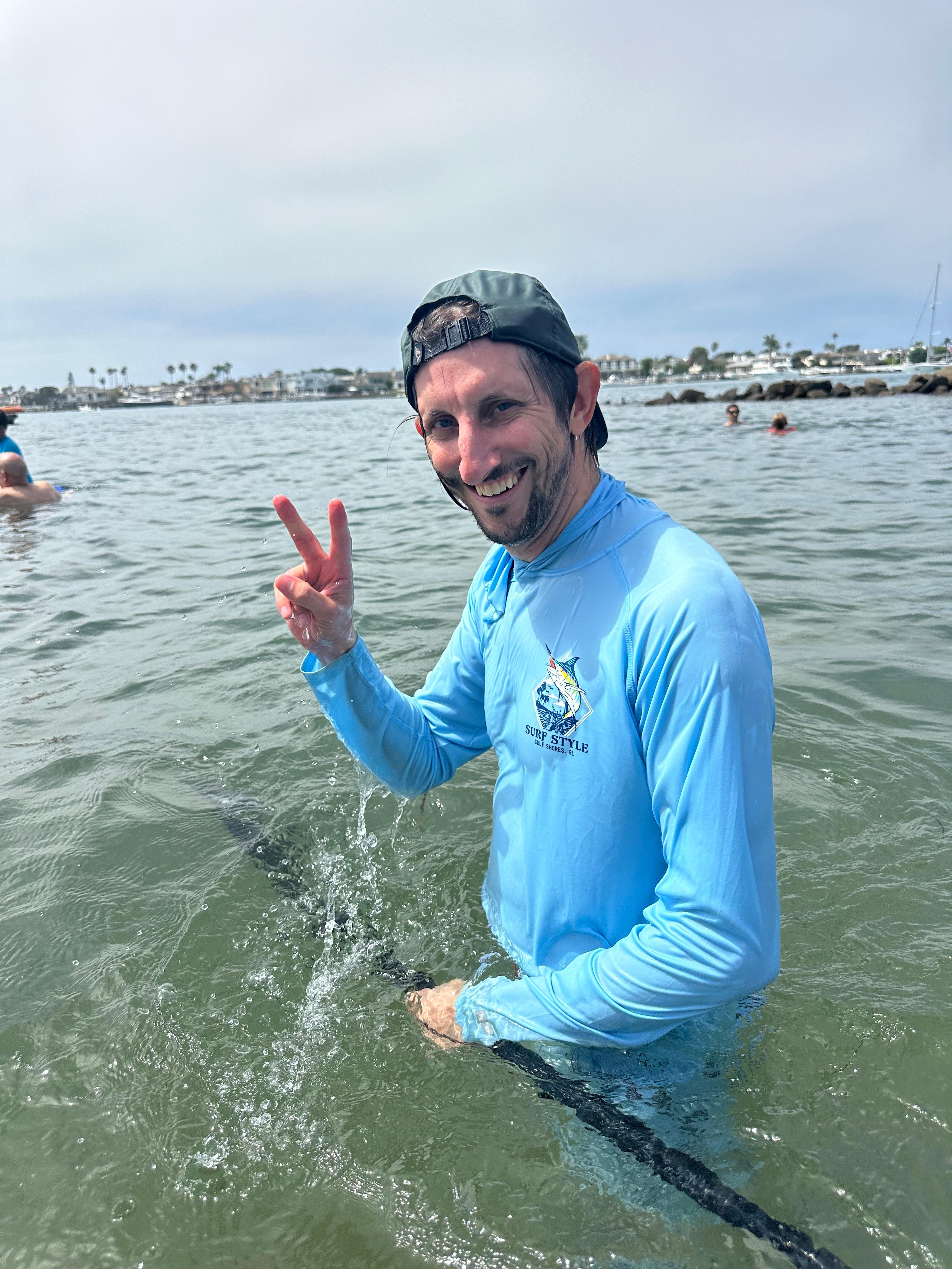 A smiling swimmer in a light-blue sun shirt flashes a peace sign while wading in a harbor, holding a rope line