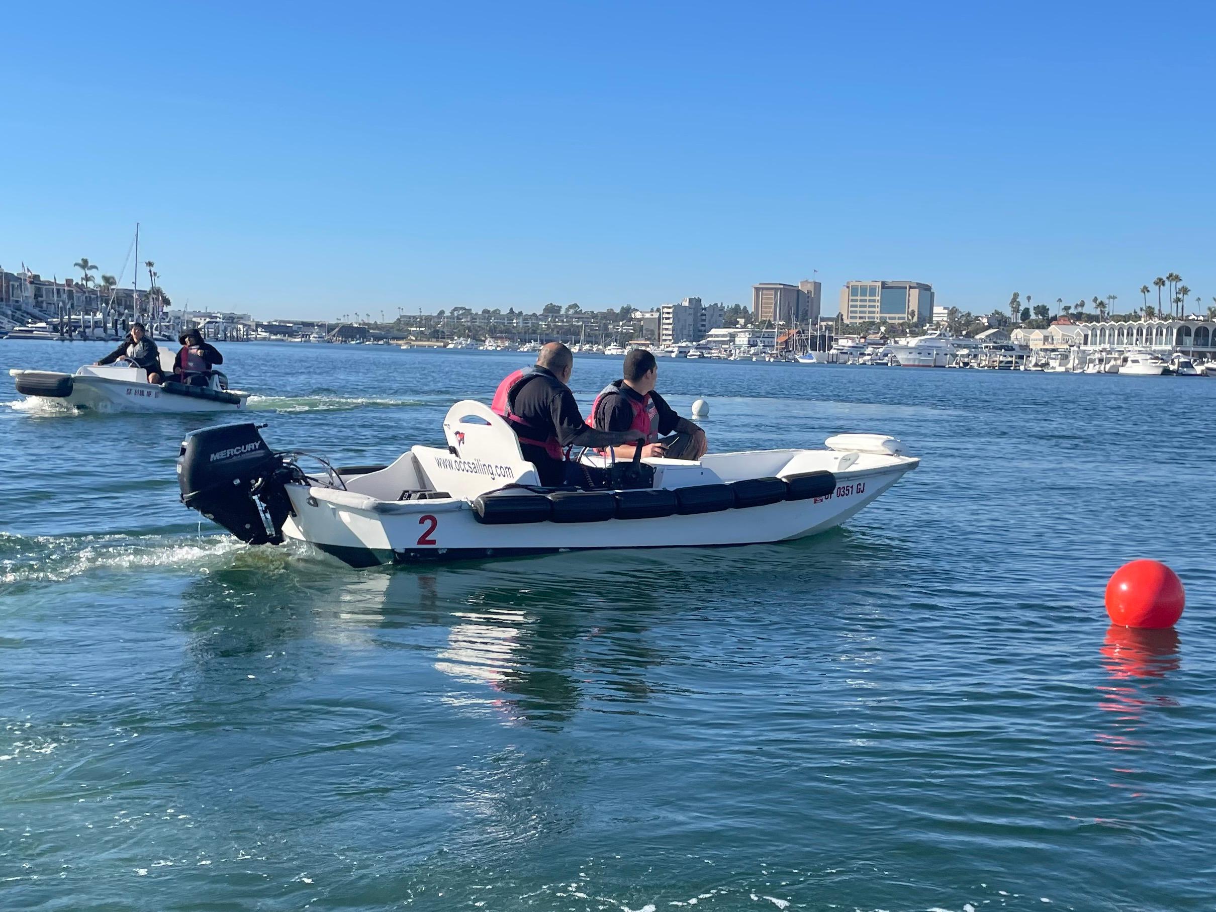 Participants in life vests drive small white training powerboats across Newport Harbor past a red buoy, with the waterfront behind