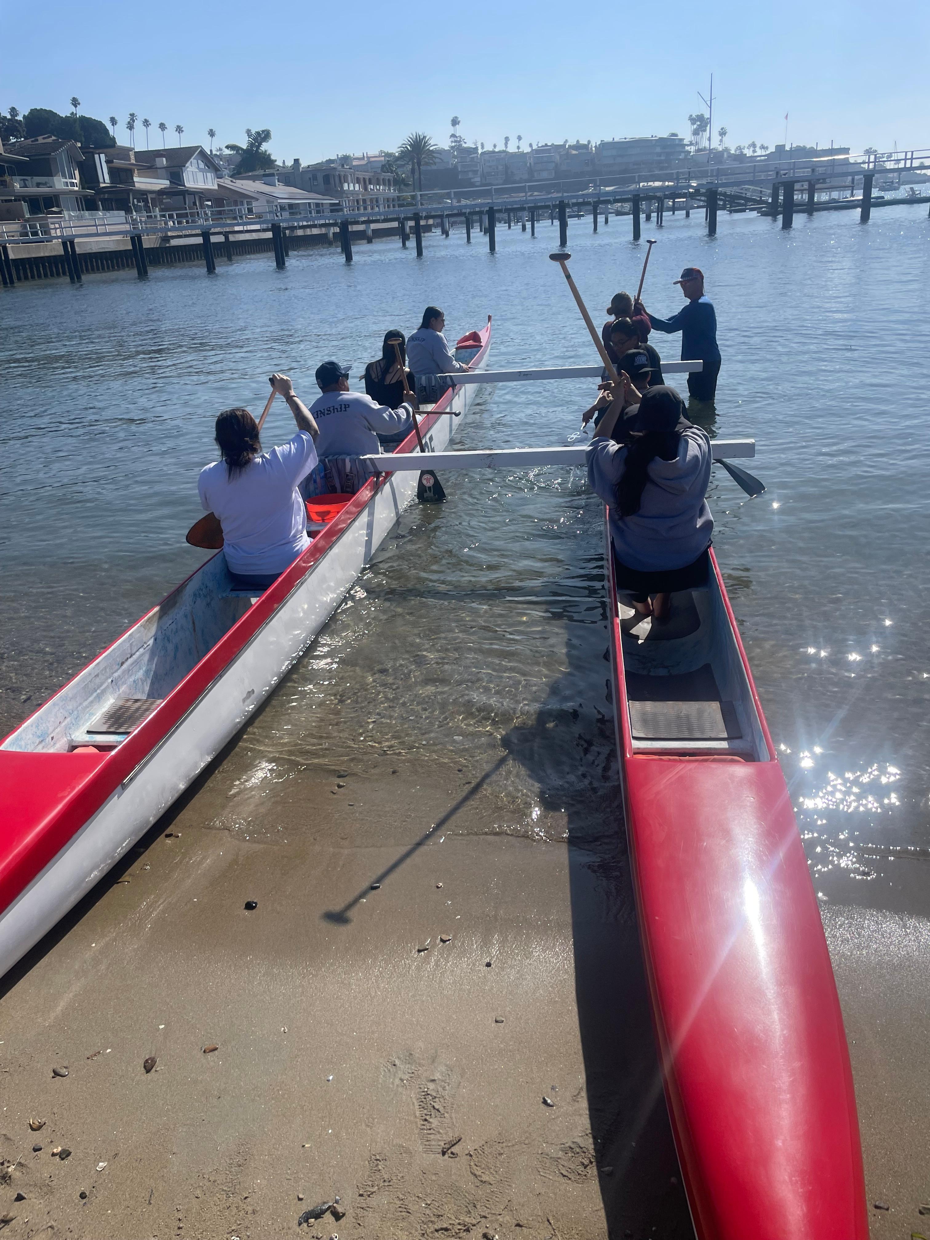 Paddlers climb into two red outrigger canoes at the edge of a calm harbor beach, with docks and waterfront homes behind