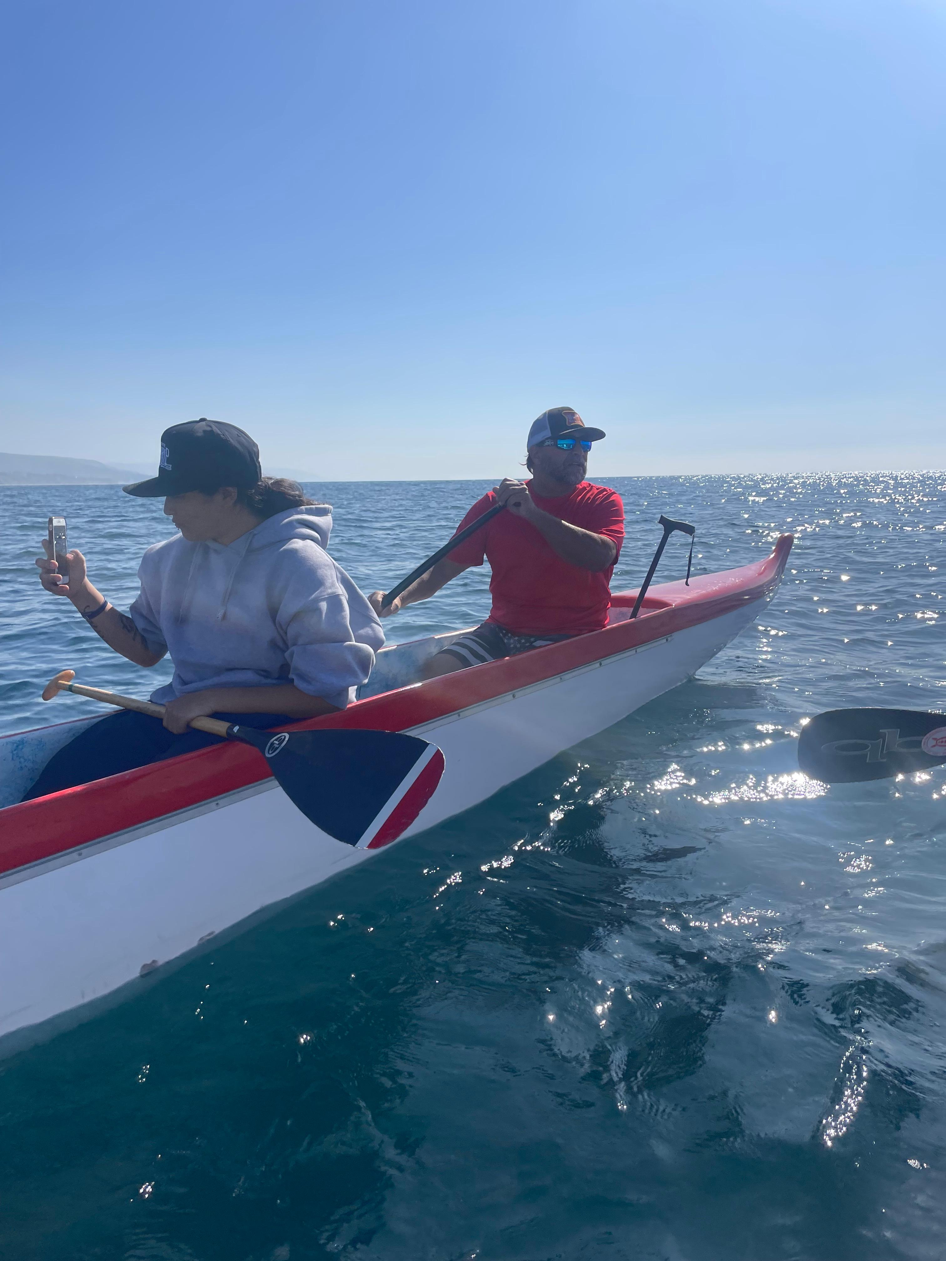 Two paddlers in a red-and-white outrigger canoe on a calm, sunlit ocean