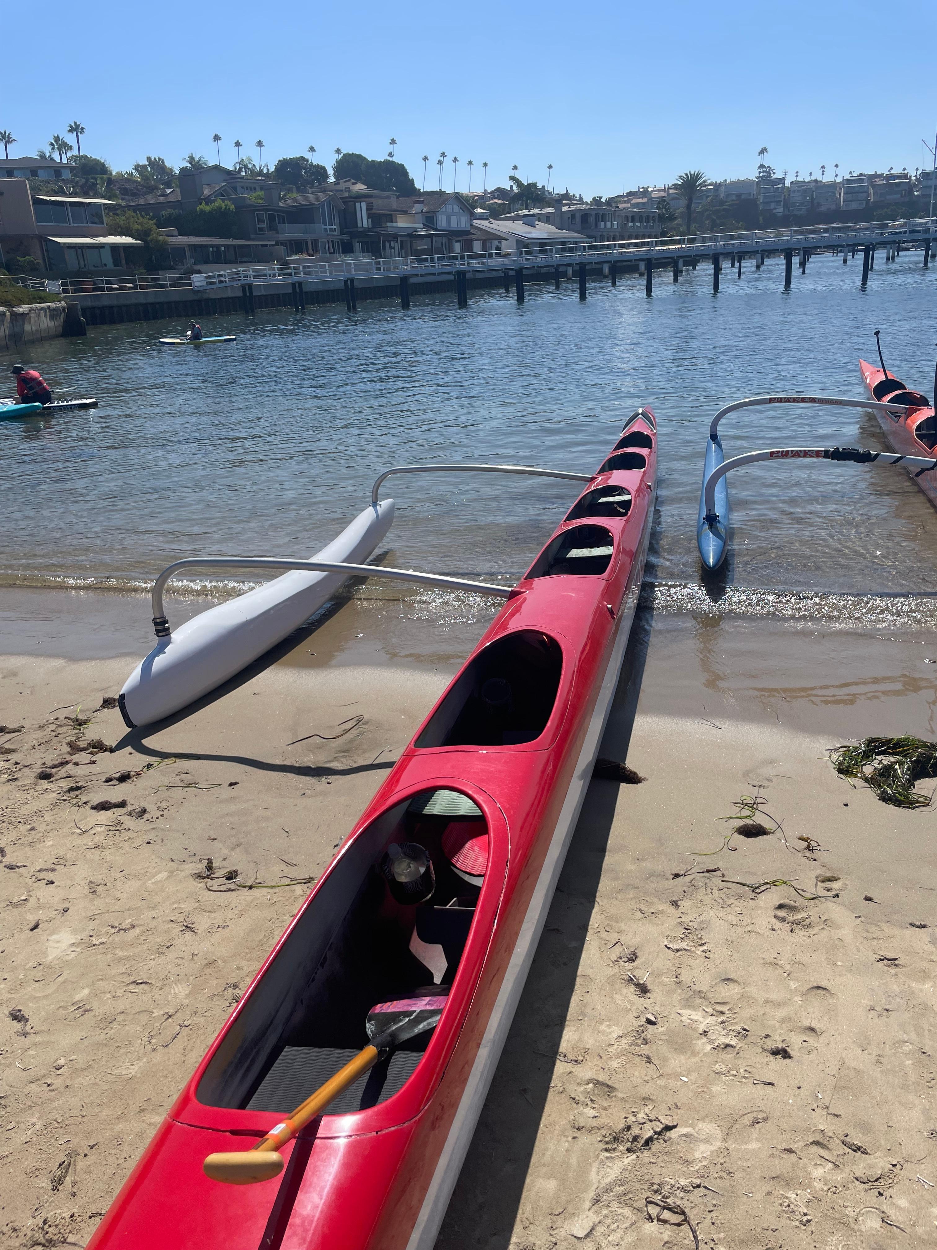Red outrigger canoes pulled up on a sandy harbor beach, with paddleboarders on the water behind