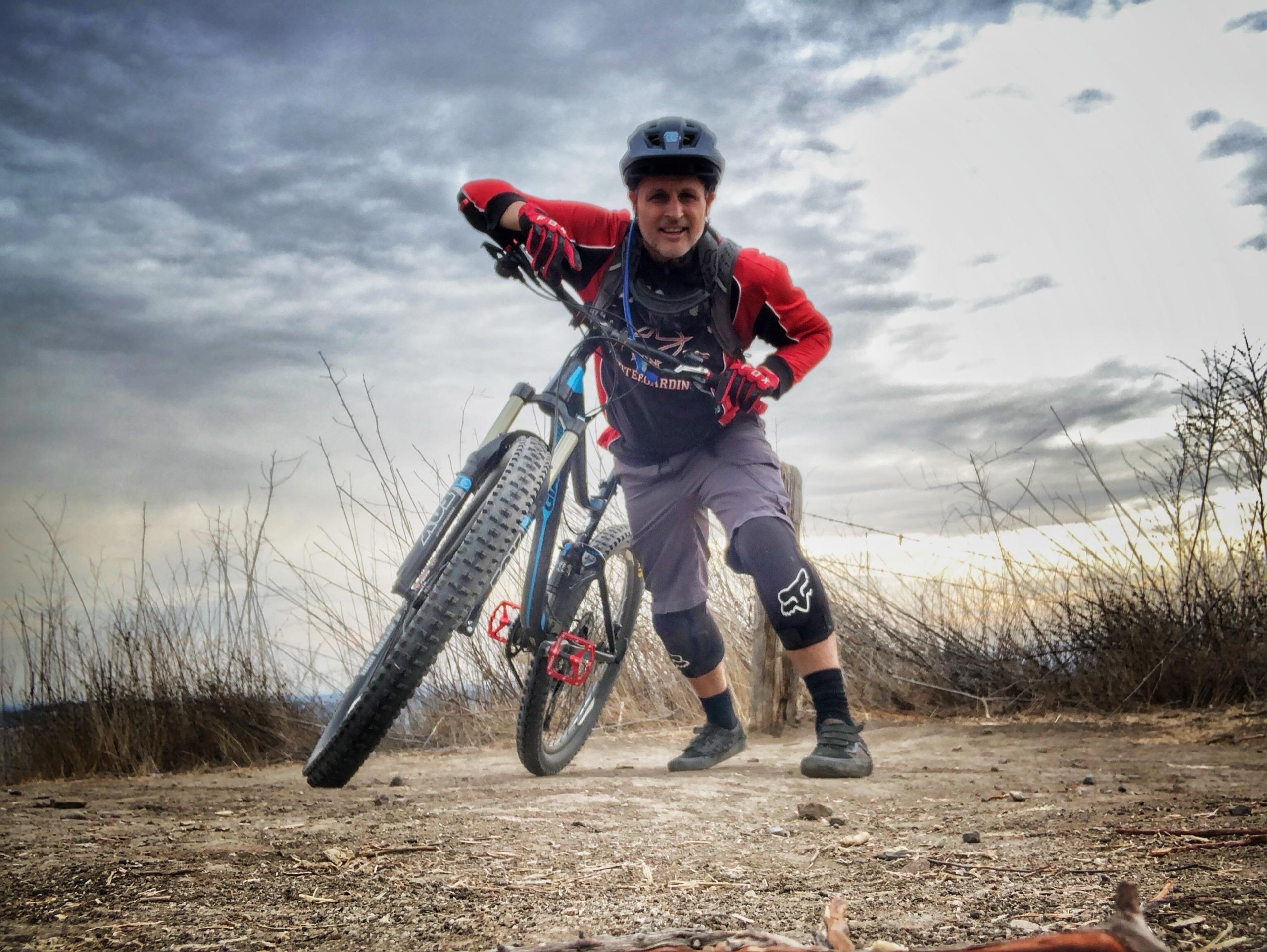 A smiling mountain biker in a helmet and red jersey leans over his bike on a dirt ridge trail under a cloudy sky