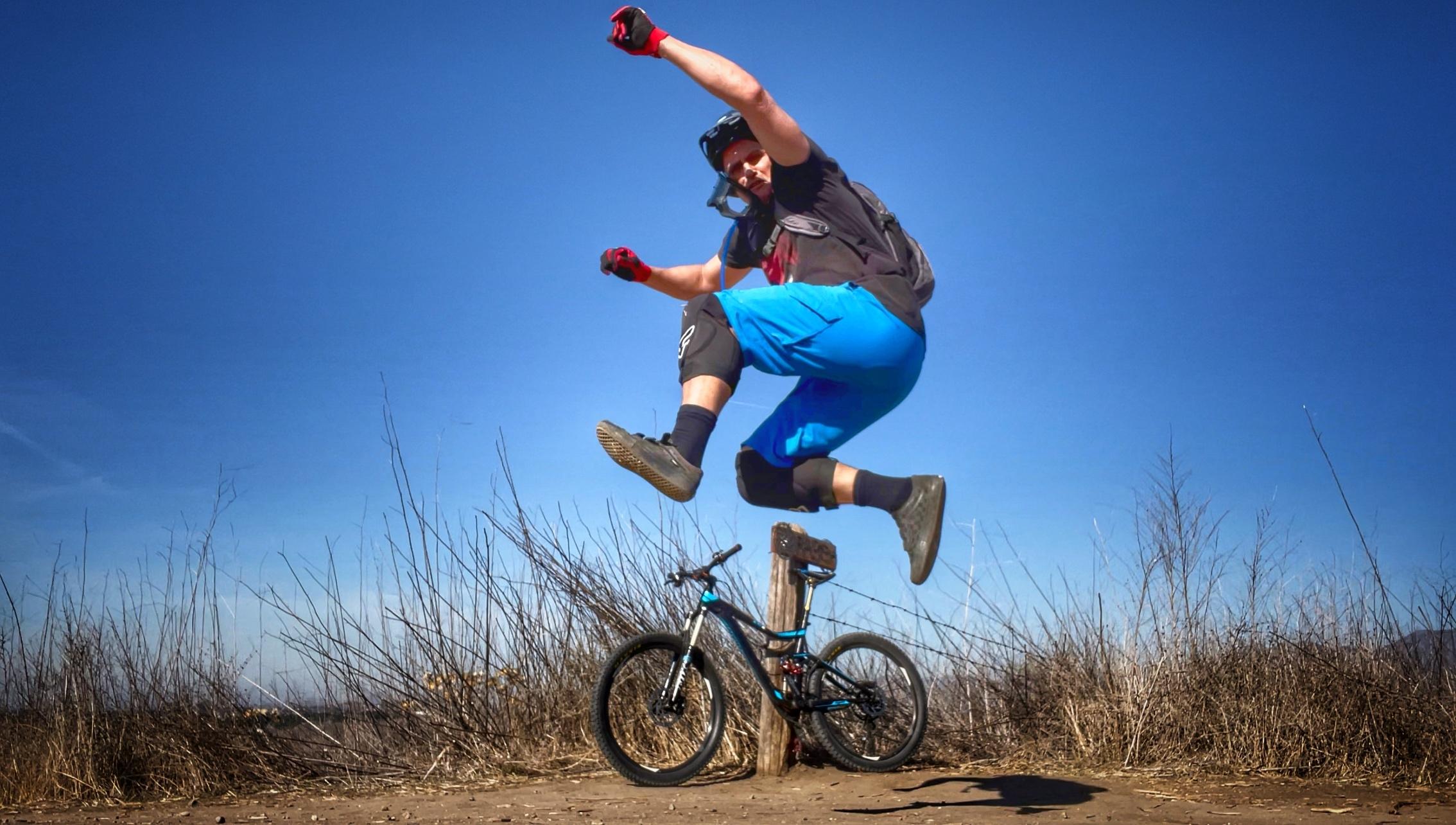 A mountain biker in full gear leaps into the air beside a bike leaning on a trail marker post