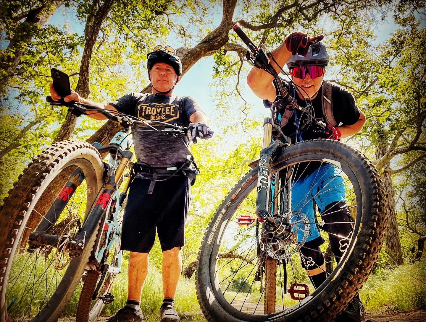 Two mountain bikers in helmets pose with their bikes under oak trees on a shaded trail